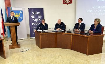 Zdjęcie przedstawiające półokrągły stuł snajdujący się na sali konferencyjnej. Przy stole siedzą cztery osoby.Od lewej dwóch policjantów, męższczyzna w garniturze oraz kobueta w błękitnym żakiecie. Z lewej strony z mówincy przemawia ubrany w garnitur mężczyzna. Za nim widać dwie plansze z logo z herbem powiatu oraz policji